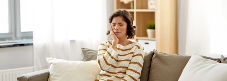 Woman on her couch holding her cheek in pain, wondering if her tooth pain is a dental emergency